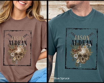 May include: Two t-shirts with a distressed graphic design featuring a skull with flowers and butterflies. The text "Jason Aldean" is printed above the skull. The t-shirts are available in Espresso and Blue Spruce.