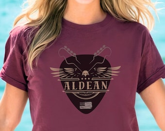 May include: A maroon t-shirt with a graphic of a guitar pick with an eagle and the text "ALDEAN" in a vintage style.