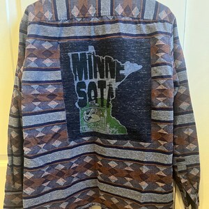 May include: A blue, brown, and gray patterned flannel shirt with a patch on the back that says "Minnesota" with an outline of the state and a cartoon character.