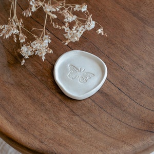 May include: A white ceramic trinket dish with a raised butterfly design. The dish is on a wooden surface.
