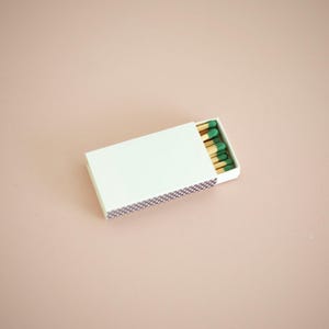 May include: A white matchbox, partially open, revealing several matches with green tips. The box is rectangular and has a purple and white patterned striking surface. The background is a solid, light pink color.