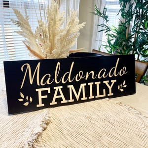 May include: Black wooden sign with gold lettering that reads "Maldonado FAMILY" with a small gold leaf design on either side of the text.