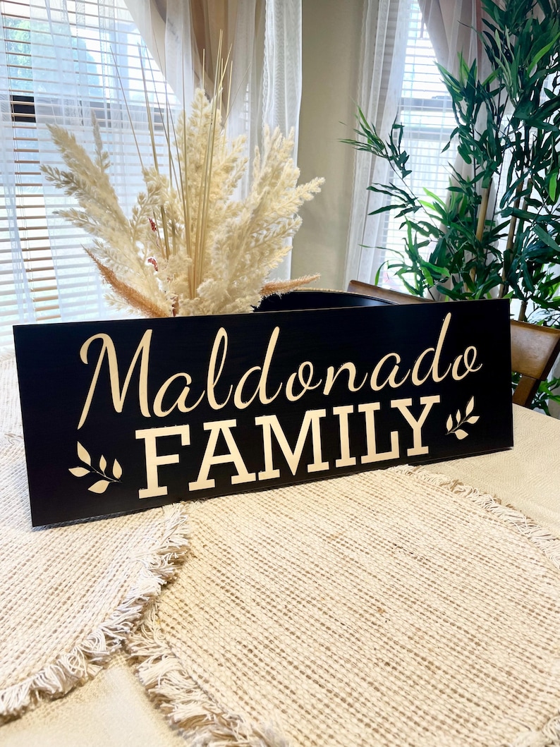 May include: Black wooden sign with gold lettering that reads "Maldonado FAMILY" with a small gold leaf design on either side of the text.