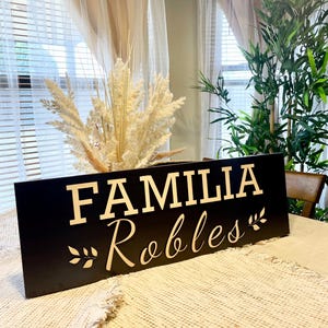 May include: Black wooden sign with the text "FAMILIA Robles" in gold lettering. The sign has a decorative leaf design on either side of the text.