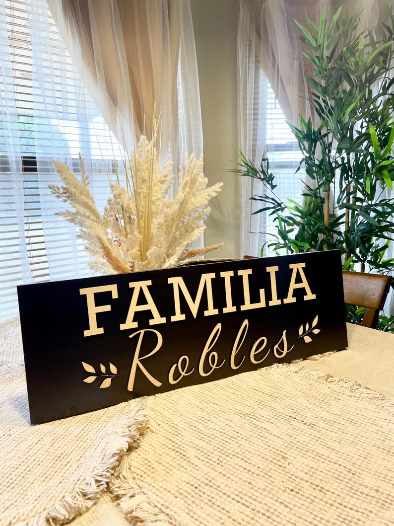 May include: Black wooden sign with the text "FAMILIA Robles" in gold lettering. The sign has a decorative leaf design on either side of the text.