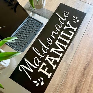 May include: Black wooden sign with white lettering that reads "Maldonado Family" with a leafy design on either side of the text.