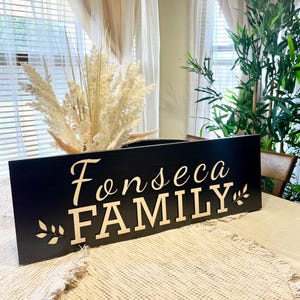 May include: Black wooden sign with the text "Fonseca FAMILY" in gold lettering. The sign is decorated with a small leaf design on either side of the text.