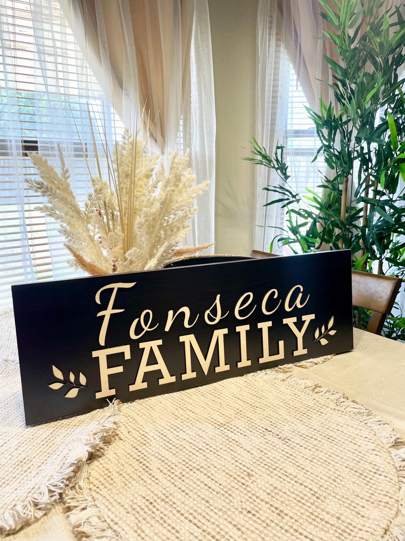 May include: Black wooden sign with the text "Fonseca FAMILY" in gold lettering. The sign is decorated with a small leaf design on either side of the text.