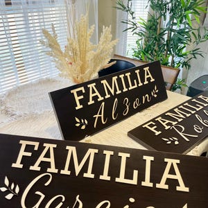 May include: Three wooden signs with the words "Familia Alzen", "Familia Robles", and "Familia Garcia" in white lettering on a dark brown background. The signs are decorated with small leafy sprigs.