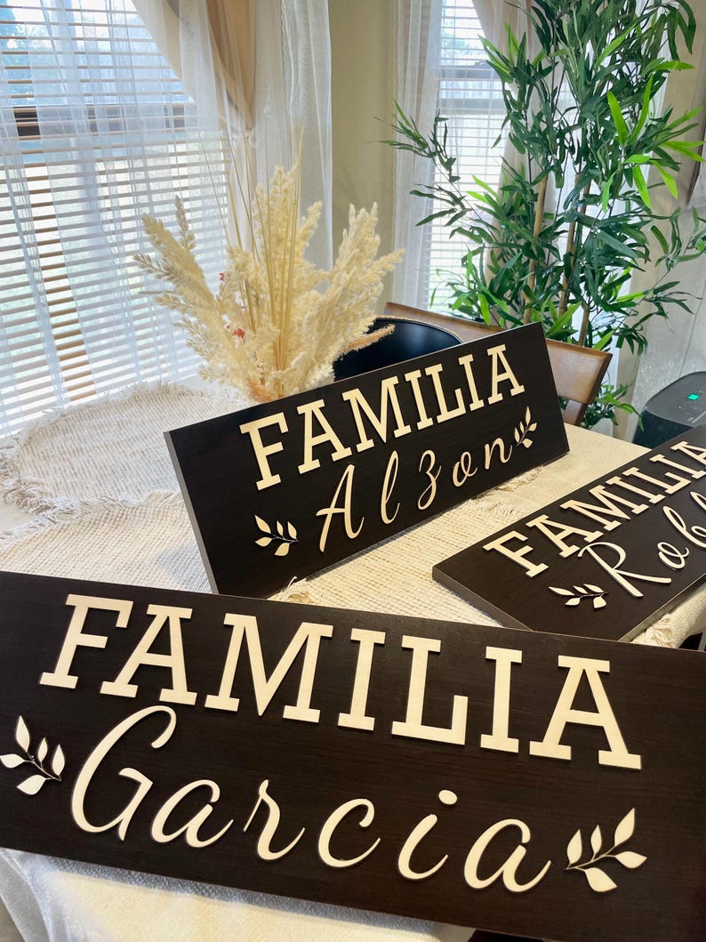 May include: Three wooden signs with the words "Familia Alzen", "Familia Robles", and "Familia Garcia" in white lettering on a dark brown background. The signs are decorated with small leafy sprigs.