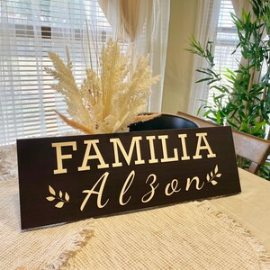 May include: A dark brown wooden sign with the words "FAMILIA Alzon" in gold lettering and a small gold leaf design on each side of the text.