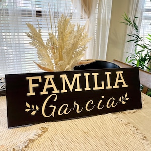 May include: A black wooden sign with the words "FAMILIA Garcia" in gold lettering and a small leafy branch on either side of the text.