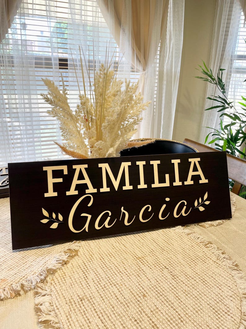 May include: A black wooden sign with the words "FAMILIA Garcia" in gold lettering and a small leafy branch on either side of the text.