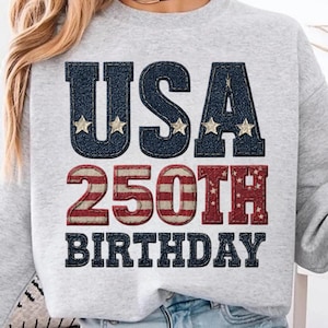 Op de afbeelding: Een lichtgrijs sweatshirt met de tekst "USA 250TH BIRTHDAY" in marineblauwe letters in denimstijl met gouden sterren en rood-witte strepen. De sweatshirt heeft een casual stijl.
