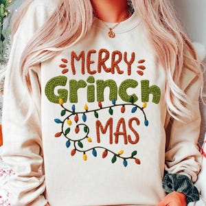 May include: Cream-colored sweatshirt with the text "MERRY Grinch MAS" in a festive design. The word "Grinch" is in green, textured letters, and the other words are in a reddish-brown color. A string of colorful Christmas lights decorates the bottom.