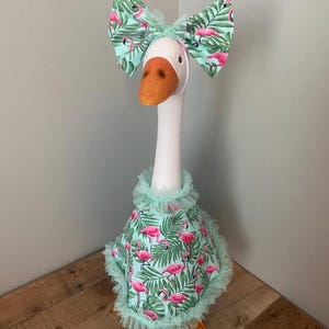 May include: A white goose wearing a teal bow with a flamingo and leaf pattern. The goose also wears a matching cape with a ruffled collar and hem. The cape features a flamingo and leaf pattern.