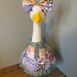 May include: A stuffed goose with a white body, yellow beak, and black eyes. It wears a patchwork patterned bow and a matching cape with green lace trim. The bow features floral and geometric patterns.
