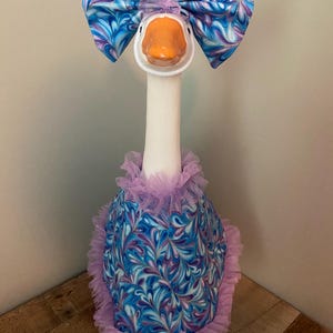May include: A decorative goose figurine with a white neck and orange beak. It is adorned with a large blue and purple patterned bow, a matching dress, and a ruffled lavender trim. The goose is a whimsical decorative item.