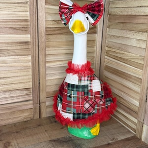 May include: A white goose statue wearing a red, green, and white plaid patchwork dress with a red tulle ruffle trim at the neck and hem. The goose also wears a matching large plaid bow on its head and has a bright yellow beak and feet.