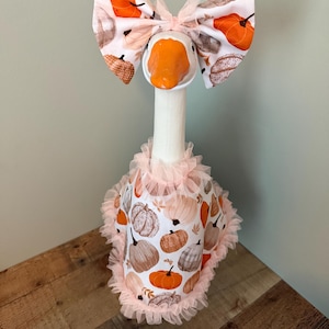May include: A decorative goose figurine wearing a fall-themed outfit. The goose has an orange beak and is adorned with a large bow featuring orange pumpkins and fall leaves. The goose is wearing a dress with a pumpkin pattern and a pink ruffled trim.