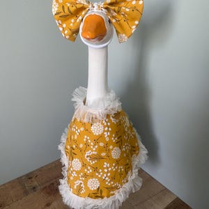 May include: A decorative goose dressed in a mustard yellow floral print bow, dress, and collar. The bow and dress feature a white floral pattern. The dress is trimmed with white lace. The goose has an orange beak.
