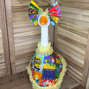 May include: A white goose figurine wearing a school-themed outfit featuring a yellow school bus, red apples, and the letters ABCD. The goose has a large, multi-colored fabric bow on its head and a yellow ruffled tulle collar and hem trim.