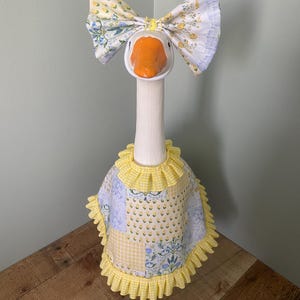 May include: A decorative goose figurine with a white neck and head, featuring an orange beak. It wears a large bow with floral patterns and a yellow-trimmed apron. The apron has a patchwork design with blue and yellow floral and gingham patterns.