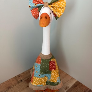 May include: A white decorative goose figurine wearing a patchwork dress and a matching oversized bow headband. The fabric features a mix of orange, yellow, green, and blue patterns with small floral and polka dot prints, finished with a tan fringe trim.