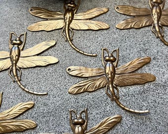 May include: Several bronze-colored dragonfly ornaments are arranged on a gray surface. Each dragonfly has detailed wings and a slender body. The ornaments are likely for crafting or decorative purposes.