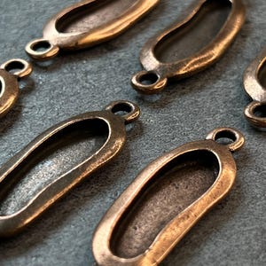 May include: Close-up of several antique bronze-colored oval connector charms. Each charm has a central elongated opening and small loops at each end. The charms are arranged on a gray surface.