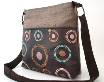 May include: A brown canvas and leather shoulder bag with a long brown strap. The bag has a flap with a brown leather pocket featuring a colorful embroidered pattern of circles.