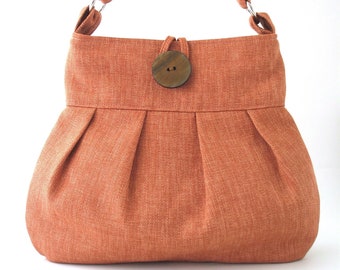 May include: A terracotta-colored canvas tote bag with a wooden button closure. The bag has a pleated design and a long shoulder strap.