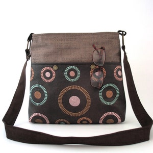 May include: A brown canvas and leather crossbody bag with a brown strap. The bag has a flap with a brown fabric top and a brown leather bottom with a circle pattern in pink, brown, and green.
