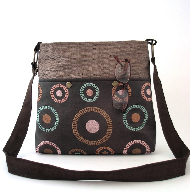 May include: A brown canvas and leather crossbody bag with a brown strap. The bag has a flap with a brown fabric top and a brown leather bottom with a circle pattern in pink, brown, and green.