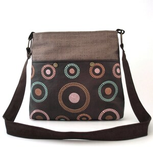 May include: A brown canvas and leather crossbody bag with a flap top and a long adjustable strap. The bag has a brown and black fabric lining with a circle pattern in pink, orange, and teal.
