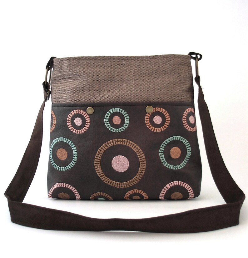 May include: A brown canvas and leather crossbody bag with a flap top and a long adjustable strap. The bag has a brown and black fabric lining with a circle pattern in pink, orange, and teal.