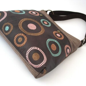 May include: A brown canvas and leather shoulder bag with a colorful circle pattern. The circles are embroidered with brown, pink, and teal thread.
