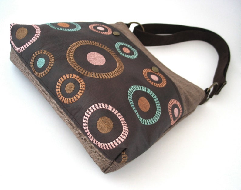 May include: A brown canvas and leather shoulder bag with a colorful circle pattern. The circles are embroidered with brown, pink, and teal thread.