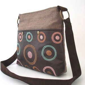 May include: A brown canvas and leather shoulder bag with a brown strap. The bag has a brown leather flap with colorful embroidered circles.