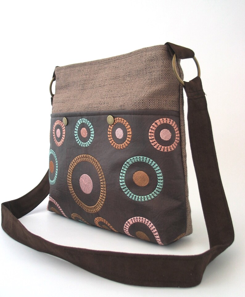 May include: A brown canvas and leather shoulder bag with a brown strap. The bag has a brown leather flap with colorful embroidered circles.