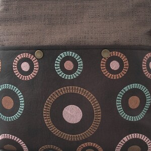 May include: A brown leather bag with a brown fabric flap and a pattern of embroidered circles in shades of pink, teal, and brown.