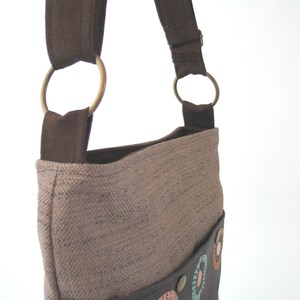 May include: A brown canvas shoulder bag with a brown leather flap and a colorful circle pattern. The bag has a brown strap with two metal rings.