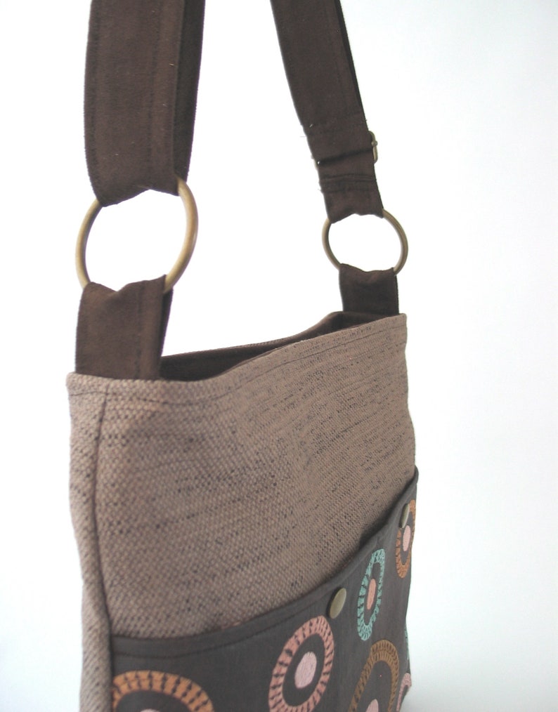 May include: A brown canvas shoulder bag with a brown leather flap and a colorful circle pattern. The bag has a brown strap with two metal rings.