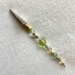 K&ouml;nnte beinhalten: Eine silberne Krokodilklemme mit goldfarbener Drahtwicklung und einem Perlenakzent. Die Perlen sind gr&uuml;n, klar und goldfarben, mit einer dekorativen Blumenperle. Der Text "Luni Loka" ist sichtbar.
