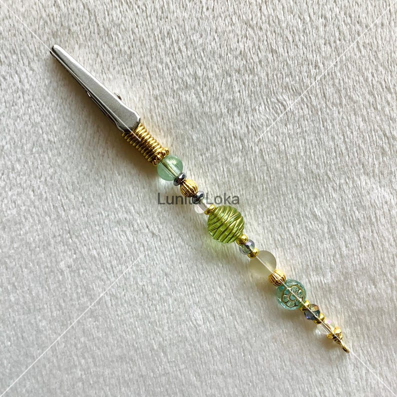 K&ouml;nnte beinhalten: Eine silberne Krokodilklemme mit goldfarbener Drahtwicklung und einem Perlenakzent. Die Perlen sind gr&uuml;n, klar und goldfarben, mit einer dekorativen Blumenperle. Der Text "Luni Loka" ist sichtbar.