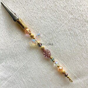 K&ouml;nnte beinhalten: Eine dekorative Krokodilklemme mit einem goldfarbenen Metallk&ouml;rper und einer Feder. Die Klemme ist mit Perlen in Rosa, Gold und Blau sowie einem Schmetterling und einem Blumenanh&auml;nger verziert. Der Text "Lunita Loka" ist sichtbar.