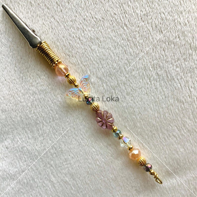 K&ouml;nnte beinhalten: Eine dekorative Krokodilklemme mit einem goldfarbenen Metallk&ouml;rper und einer Feder. Die Klemme ist mit Perlen in Rosa, Gold und Blau sowie einem Schmetterling und einem Blumenanh&auml;nger verziert. Der Text "Lunita Loka" ist sichtbar.