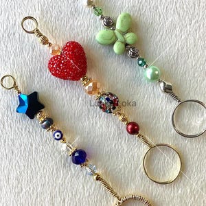 May include: Three decorative keychains with unique designs. One features a red heart, another a green butterfly, and the third a blue star. Each keychain has a gold-colored ring and is adorned with various beads and charms.