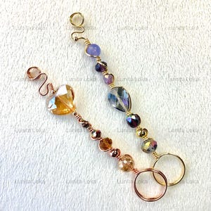 May include: Two handmade hair accessories. One features a heart-shaped amber crystal, the other has a teardrop-shaped crystal. Both have a spiral design and are adorned with various beads in shades of gold, purple, and amber. The accessories are made with gold and copper wire.