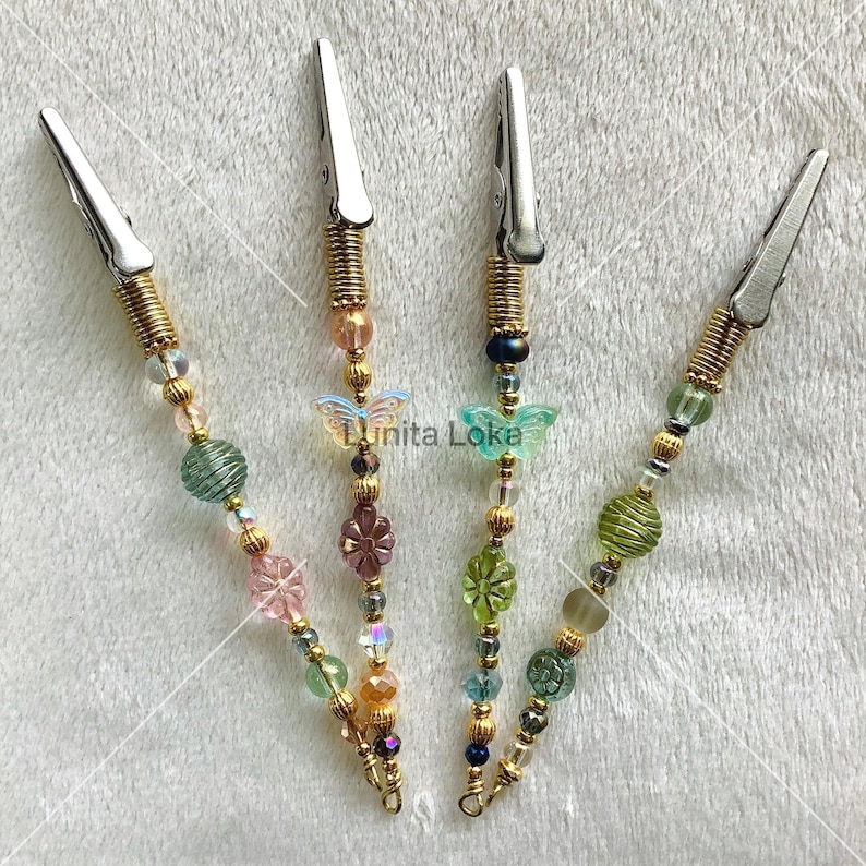 K&ouml;nnte beinhalten: Vier dekorative Metallclips mit Perlenakzenten. Jeder Clip hat einen silberfarbenen Clip, eine goldfarbene Feder und eine Kette aus bunten Perlen. Die Perlen umfassen verschiedene Formen und Farben, darunter Schmetterlinge und florale Designs.
